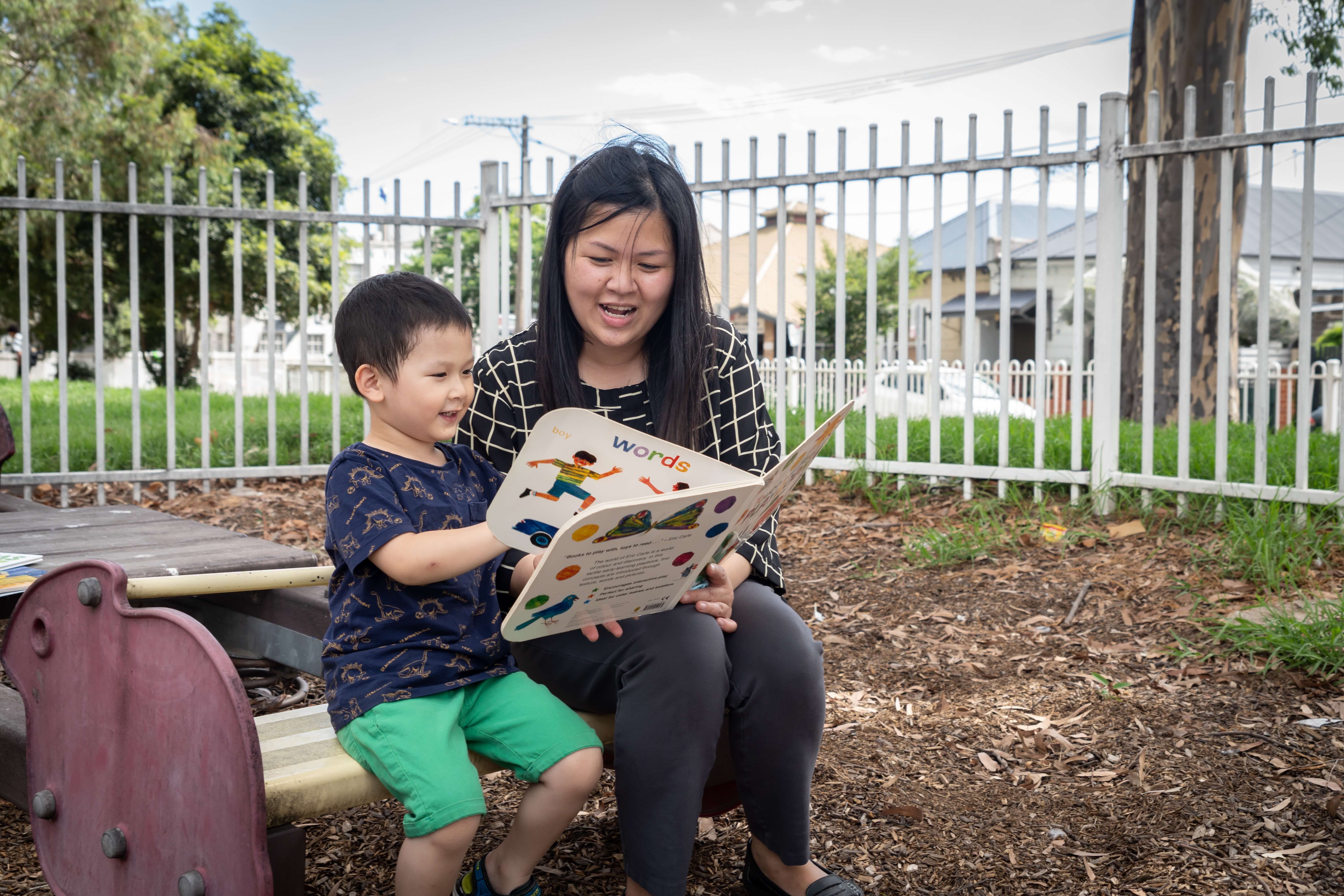 A young student reads a book with help from a teacher in an outdoor playground.